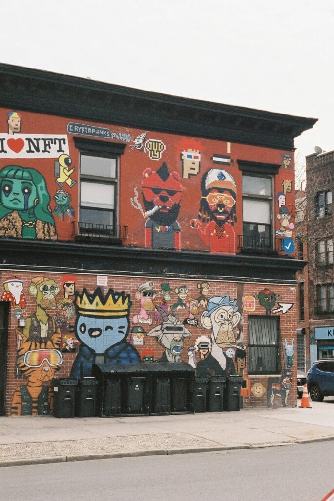 Street mural on a red-brick building featuring various NFT PFP characters, including pixel art faces, Bored Apes, and other cartoon-style figures, celebrating NFT culture.