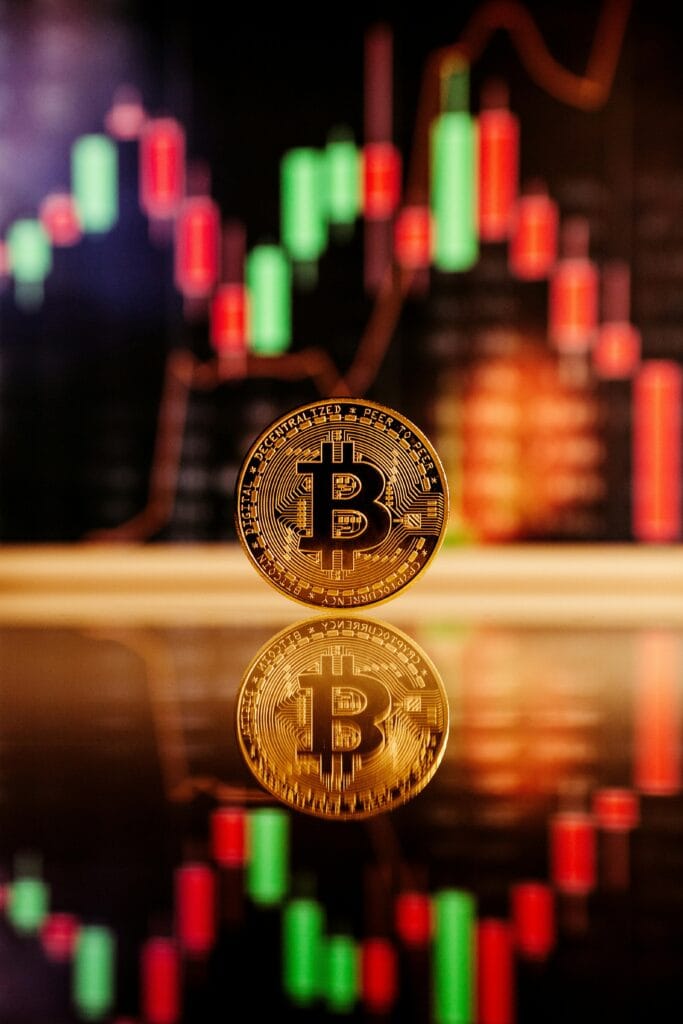 Golden Bitcoin coin standing on a reflective surface with candlestick trading charts in the blurred background.