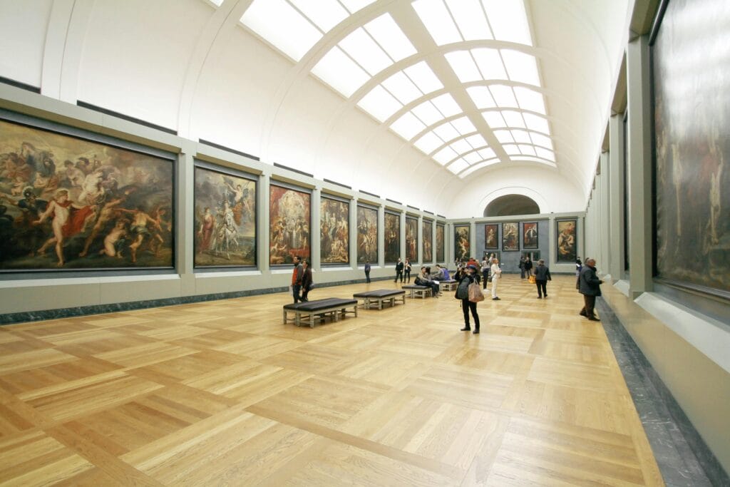 Wide museum gallery with large classical paintings on both walls, high arched ceiling with skylight, and visitors observing the artworks and walking on polished wooden floor.