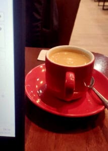 A red cup filled with coffee on a matching saucer with a spoon, placed on a wooden café table beside a laptop screen, creating a cozy morning atmosphere.