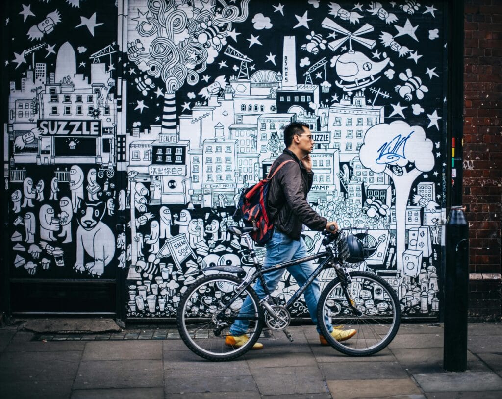 Street photo of a man walking his bicycle in front of a large black-and-white mural with surreal cityscapes, cartoon characters, and playful details. The mural reflects pop surrealism influences, connecting to Indonesian contemporary art and the practices of NFT artists, blending urban culture with experimental visual storytelling.
