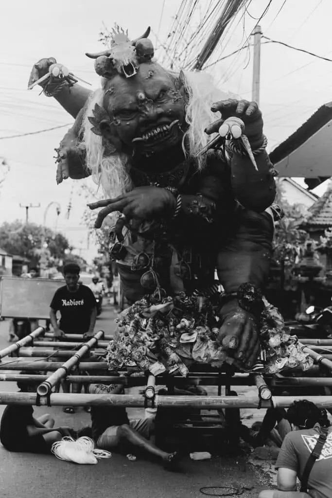 Black and white photo of a grotesque Balinese ogoh-ogoh effigy for the Nyepi festival, featuring sharp teeth, skull crown, and exaggerated monstrous features, carried on a bamboo frame by young men in a village street.
