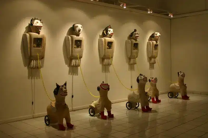 Heri Dono’s contemporary art installation Born and Freedom, featured on Artsy, showing wall-mounted winged humanoid sculptures connected by yellow chains to wheeled figures.