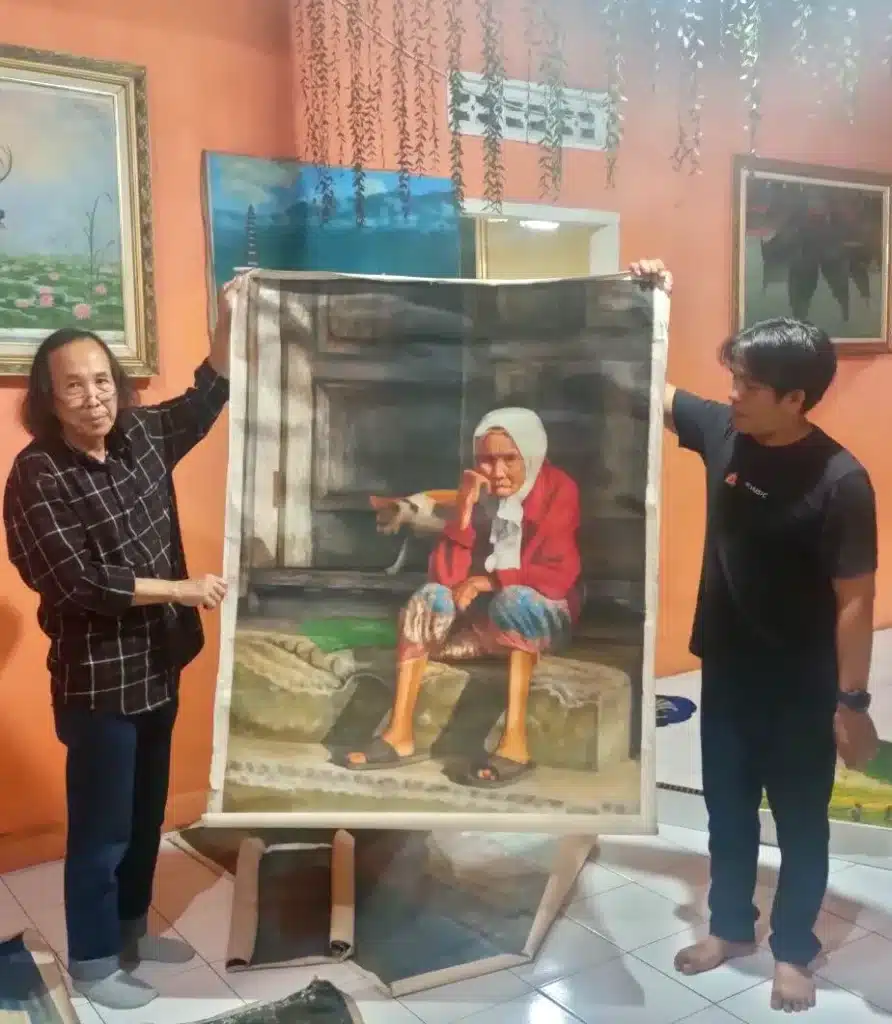 Senior Indonesian art curator Hendro Wiyanto and painter Pandji Sumantri holding up a large realism canvas painting of an elderly woman during a studio visit.